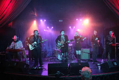 Six musicians of Roselit Bone perform under dramatic purple and amber stage lighting at Portland's iconic Dante's venue during RX Fest 2019. The intimate club setting frames the band with rich crimson curtains while atmospheric haze swirls through the colored beams, creating a moody backdrop for their eclectic instrumentation including guitars, violin, and keyboards. The low stage positioning places the audience at eye level with the performers, emphasizing the venue's legendary intimacy.