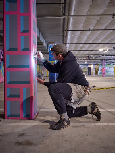 Portland bike activist and artist Rabbitt kneels in concentrated focus as he applies spray paint to a concrete pillar in the Clay Creative building's parking garage. His geometric composition features bold pink and blue rectangular forms that contrast sharply against the raw industrial architecture. The underground space, illuminated by harsh fluorescent lighting, transforms into an impromptu studio where urban art meets utilitarian infrastructure.