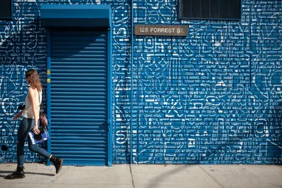 A woman in casual attire strides purposefully past an extraordinary blue mural that transforms the entire facade of 128 Forrest Street in Brooklyn into a mesmerizing maze of white geometric patterns and symbols. The monochromatic artwork creates an optical illusion of depth and movement, with intricate line work covering every surface from the roll-up security gate to the surrounding walls. The pedestrian's natural movement provides a striking human counterpoint to the static yet visually dynamic street art, while bright daylight enhances the vivid cobalt blue that dominates the urban canvas.