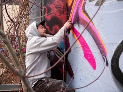 Travis Fields, known as Campographic, meticulously applies vibrant magenta and orange spray paint to a concrete wall at the Clay Creative Building in Portland's Hosford-Abernethy neighborhood. Captured mid-creation, the hooded artist works with focused intensity among dormant winter branches, transforming the urban canvas with bold geometric patterns. The industrial setting frames this moment of artistic genesis, where raw concrete surrenders to explosive color.