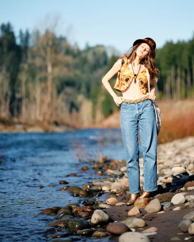 A young woman with flowing brown hair tilts her head back in peaceful contemplation, standing barefoot on the rocky riverbed of Oregon's Floodplain Trail. Bathed in warm golden light, she wears a fox-print crop top, high-waisted jeans, and a dark fedora, her eyes closed in serene meditation. The Columbia River's gentle waters lap against smooth river stones while autumn trees blur softly in the background, creating an intimate moment of connection with the Pacific Northwest wilderness.