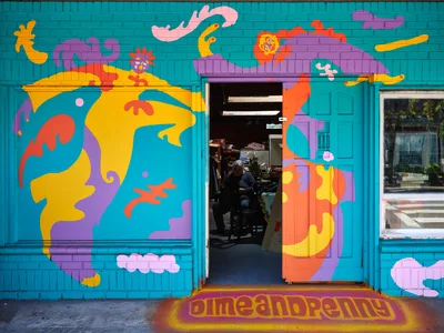 Artist Naomi Likayi's explosive mural transforms a turquoise storefront in Portland's Alberta Arts District into a portal of liquid imagination. Swirling forms in golden yellow, coral orange, and violet purple cascade across the facade like molten consciousness, while the open doorway reveals glimpses of the establishment's interior. The psychedelic composition pulses with organic energy against the structured geometry of painted brick, creating an inviting threshold between the everyday sidewalk and a realm of unbridled creativity.