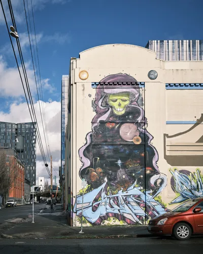 A towering street mural transforms the curved facade of a former Salvation Army warehouse on Southeast Ash Street in Portland's Central Eastside district. The artwork depicts a haunting lime-green skull wreathed in purple cosmic smoke, its hollow sockets gazing over a star-filled cosmic landscape populated with planets and celestial debris. Power lines slice diagonally through the bright blue afternoon sky, while modern apartment buildings frame the scene and a red sedan parks casually beneath this monumental display of urban art.