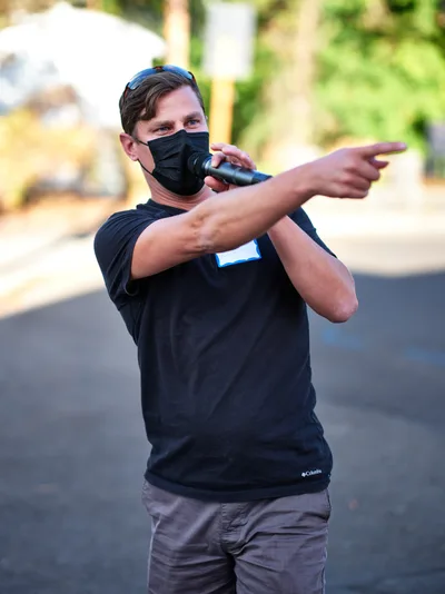 Michael Gaskill, wearing a black face mask and Columbia shirt, gestures emphatically while speaking into a microphone during a demonstration outside Representative Kurt Schrader's Oregon City office. Golden afternoon light filters through the trees behind him, creating a warm bokeh backdrop as he addresses the assembled crowd on McLoughlin Promenade. His animated stance and pointing gesture convey passionate advocacy, captured in this moment of civic engagement.