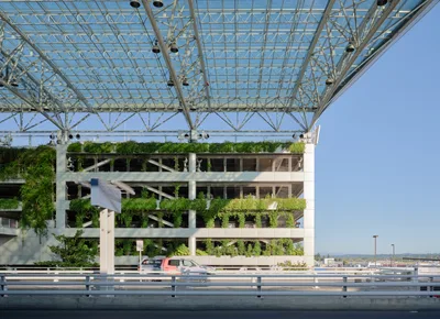 A striking view of Portland International Airport's innovative short-term parking garage showcases the marriage of sustainable design and modern infrastructure. Cascading vegetation transforms the concrete structure into a verdant vertical garden, while an intricate steel and glass canopy overhead creates geometric patterns against the clear Oregon sky. The foreground highway barrier frames this architectural achievement, where a lone red vehicle provides scale to the ambitious green building initiative that defines PDX's commitment to environmental stewardship.