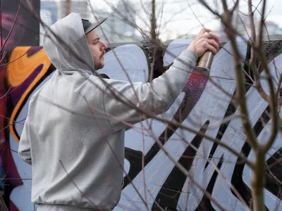 Travis Fields, known as Campographic, concentrates intently as he applies paint to a weathered concrete wall at the Clay Creative Building in southeast Portland's Hosford-Abernethy neighborhood. Clad in a gray hoodie and black cap, the muralist works with deliberate precision while bare winter branches frame the foreground, creating natural geometry against the urban canvas. The soft daylight illuminates his focused expression and the textured surface where vibrant colors begin to emerge against the industrial backdrop.
