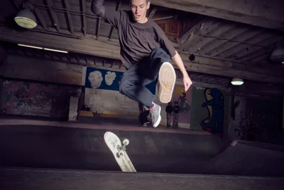 A young skateboarder executes a dynamic trick in the shadowy depths of Commonwealth Skateboarding's concrete bowl in Portland's Buckman neighborhood. The industrial ceiling beams and graffitied walls create a gritty underground atmosphere, while moody lighting illuminates the skater's focused expression mid-maneuver. The worn concrete transitions and scattered deck below speak to countless sessions in this legendary Southeast Portland skate haven.