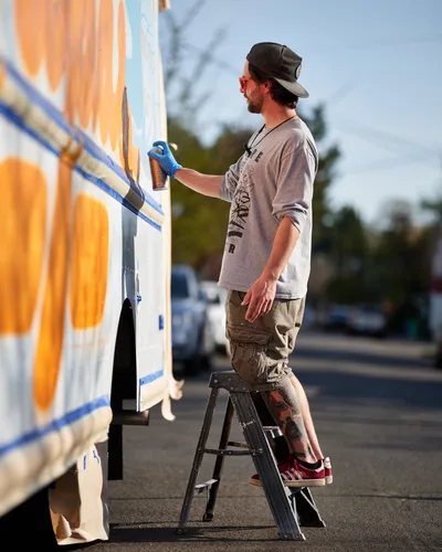 Golden hour light bathes a bearded muralist as he carefully applies spray paint to the side of a white food truck on Northeast 9th Avenue in Portland, Oregon. Standing on a weathered stepladder, the artist wears a black cap and gray long-sleeve shirt while working on vibrant orange and blue geometric designs. The warm evening light creates a luminous contrast against the emerging artwork, capturing the intersection of mobile commerce and street art culture in this King neighborhood scene.