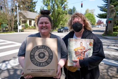 Two friends stand proudly outside Music Millennium on East Burnside Street during Record Store Day 2022, clutching their vinyl discoveries against the backdrop of Portland's leafy Kerns neighborhood. The woman on the left beams with joy while holding a Music Millennium shopping bag, her dark hair twisted into a casual top knot. Her companion, wearing a black mask and hoodie, displays a vintage Shannon and The Clams album cover, their curly blonde hair catching the filtered spring sunlight. Behind them, the quiet residential street stretches toward a traffic light, lined with mature trees and characteristic Portland homes.