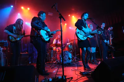 Roselit Bone commands the intimate stage at Dante's in Portland, bathed in crimson and azure stage lights that pierce through the venue's signature darkness. The band's frontwoman grips her white Gretsch guitar while delivering vocals into the microphone, flanked by leather-clad bandmates wielding electric guitars and bass. Behind them, the drummer maintains rhythm beneath a geometric kick drum design, while the warm glow of amber spotlights creates dramatic silhouettes against the club's moody atmosphere.