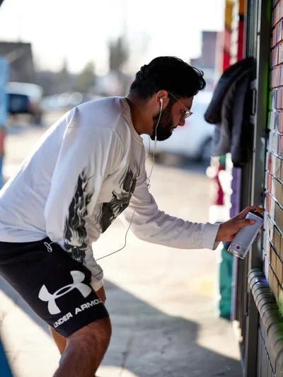 Muralist Heysus leans into his work along a vibrant wall at the intersection of Killingsworth and Northeast 15th Avenue in Portland's Alberta neighborhood. Bathed in warm afternoon light, the bearded artist wearing a white graphic long-sleeve shirt and black athletic shorts carefully selects from an array of spray paint cans, his white earbuds connecting him to his creative process. The urban streetscape behind him blurs into soft bokeh, while colorful building facades hint at the artistic energy that defines this gentrifying corner of Northeast Portland.