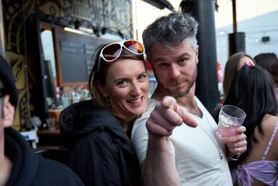 A jubilant couple shares an intimate moment at White Owl Social Club during Your Sunday Best, a popular summer day party in Portland, Oregon. The woman, wearing sunglasses and a black jacket, beams with joy as the bearded man in a cream henley playfully points toward the camera while holding a drink. The bustling venue atmosphere buzzes with conversation and laughter, captured in the warm, natural light filtering through the social club's windows.