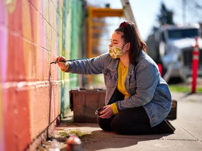 Multidisciplinary artist Vilatose sits cross-legged on the sidewalk, painting delicate details on her vibrant mural 'Jardín Cubana' depicting three female figures along Northeast Alberta Street in Portland's historic arts district. Wearing a patterned face mask and denim jacket over a golden yellow sweater, she extends her brush toward the rainbow-hued wall in warm afternoon light. The urban scene captures the intimate process of community art-making, with colorful buildings and parked vehicles softly blurred in the background.