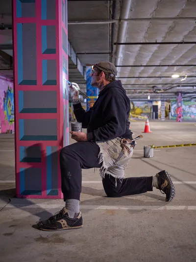 Portland artist and bike activist Rabbitt kneels in focused concentration as he meticulously applies paint to a vibrant geometric mural on a concrete pillar within the Clay Creative building's parking garage. His signature style emerges in bold pink and blue geometric patterns against the industrial backdrop of exposed ceiling beams and concrete surfaces. The intimate moment captures the transformation of urban infrastructure into artistic expression, with paint cans and brushes scattered nearby as tools of creative rebellion.