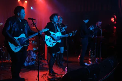 Victor Franco grips his Telecaster while Charlotte McCaslin commands center stage with her hollow-body guitar, Faith Grossnicklaus anchoring the rhythm section as Roselit Bone delivers an electrifying performance. The intimate venue pulses with crimson and azure stage lights, creating dramatic silhouettes against the smoky atmosphere of Portland's legendary Dante's. Microphone stands lean into the musical conversation while cables snake across the darkened stage, capturing the raw energy of RX Fest 2019.