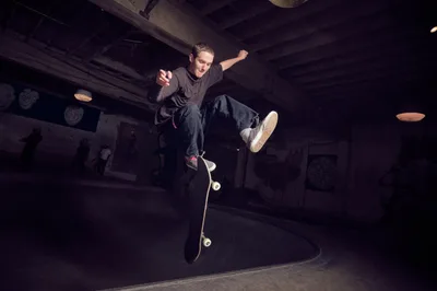 A young skateboarder launches into a dynamic kickflip above the concrete bowl at Commonwealth Skateboarding in Portland's Buckman neighborhood. The dramatic low-angle perspective captures him suspended mid-trick against the shadowy industrial ceiling, his dark clothing silhouetted by moody artificial lighting. The weathered concrete transitions and exposed beams create an authentic underground atmosphere that defines Portland's skateboarding culture.