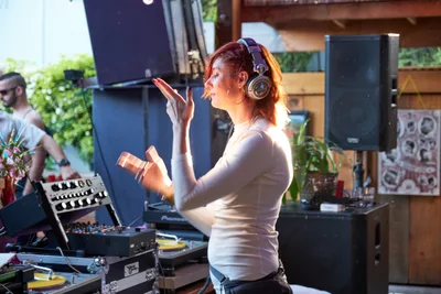 DJ Doctor Dakar commands the decks at White Owl Social Club during the Your Sunday Best season opener, her arms raised in animated gesture as she curates the afternoon's sonic journey. Golden sunlight streams through the venue's windows, illuminating her focused expression beneath professional headphones while vinyl records and synthesizer equipment create an intimate electronic music sanctuary. The Portland day party atmosphere buzzes with creative energy as patrons gather in the background among lush greenery and industrial-chic architecture.