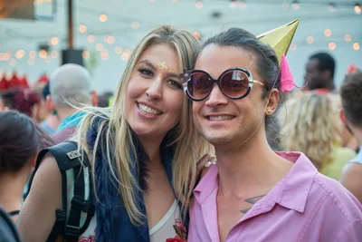 Two young women embrace with radiant smiles at Your Sunday Best day party in Portland's White Owl Social Club. The blonde woman on the left sparkles with golden glitter adorning her face, while her brunette companion sports oversized round sunglasses and a whimsical party hat with pink tassel. Behind them, the warm bokeh of string lights creates an intimate festival atmosphere as other revelers dance in the softly blurred background.