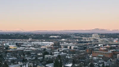 The sprawling neighborhoods of Northwest Portland unfold beneath a pastel dusk sky, where residential homes nestle between modern apartment complexes and commercial buildings. The iconic white arches of the Fremont Bridge span the distant Willamette River, while the snow-capped silhouette of Mount Saint Helens anchors the horizon in Washington State. Golden hour light bathes the urban landscape in warm tones, creating a serene contrast between the intimate scale of residential streets and the grandeur of the Cascade Range beyond.