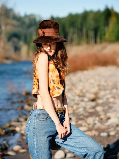 A young woman with flowing brunette hair poses confidently along the rocky riverbed of an Oregon river, wearing a distinctive brown felt hat with leather band, vintage-inspired floral crop top, and high-waisted denim. The golden hour light creates warm shadows across her face while the Columbia River Gorge's forested hillsides provide a dreamy backdrop. Her relaxed stance and gentle smile capture a moment of carefree summer exploration along the river bed.