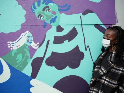 Artist Naomi Likayi stands beside her vibrant mural painted on the boarded Portland World Trade Center, wearing a white face mask and patterned coat. The artwork features flowing figures rendered in soft blues, purples, and mint greens against a rich purple background, creating a dreamlike narrative across the temporary urban canvas. Her contemplative gaze and proximity to the work establishes an intimate connection between creator and creation. The street art transforms the utilitarian boarding into a window of imagination and color.