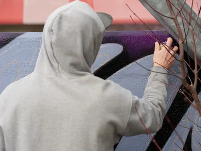 Travis Fields, known as Campographic, works intently on a vibrant mural at the Clay Creative Building in southeast Portland's Hosford-Abernethy neighborhood. Wearing a gray hoodie, the artist reaches toward purple and black paint strokes while delicate bare branches frame the foreground. The soft afternoon light illuminates the textured wall surface, capturing the meditative focus of artistic creation in this converted industrial arts space.