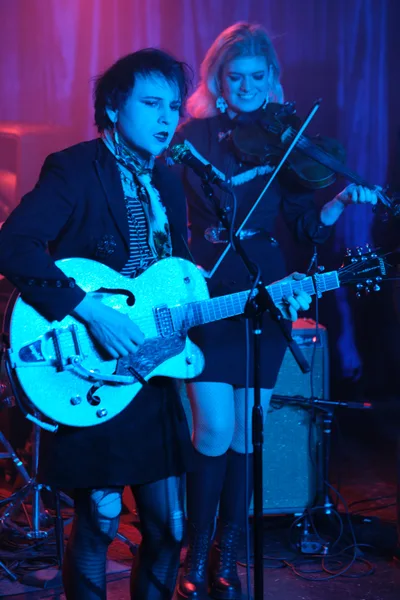 Charlotte McCaslin grips her white hollow-body guitar with fierce concentration while Faith Grossnicklaus draws her violin bow behind her, both performers bathed in the saturated purple and blue stage lights of Portland's legendary Dante's venue. The intimate underground atmosphere crackles with electric energy as the duo delivers their signature blend of rock and classical instrumentation. McCaslin's dark tousled hair and vintage-inspired outfit contrast sharply with the ethereal glow of the stage lighting, while Grossnicklaus remains partially obscured in the moody shadows.