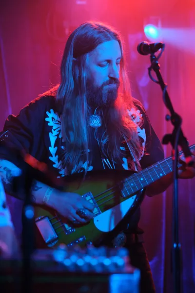 Tobias Berblinger of Roselit Bone emerges from a dreamscape of magenta and cyan stage lighting, his flowing blonde hair and full beard catching the electric glow as he cradles his bass guitar. The intimate venue pulses with atmospheric haze, transforming the musician into a spectral figure lost in concentration. His dark shirt adorned with white botanical prints creates striking contrast against the saturated colors washing over Dante's legendary stage in Portland's Old Town.