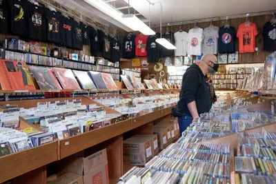 A bearded man in a black hoodie and face mask browses through wooden crates overflowing with vinyl records at Music Millennium on Burnside Street during Record Store Day 2022. The warm fluorescent lighting illuminates the cramped aisles lined with thousands of albums, creating an intimate atmosphere of musical discovery. Band t-shirts hang overhead like colorful flags, while shelves stretch toward exposed ceiling beams packed with decades of recorded music history.