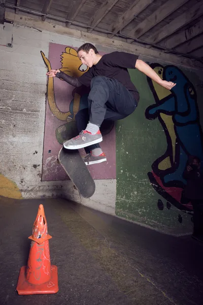A skateboarder captures air against a vibrant graffiti-covered wall in the underground confines of Commonwealth Skateboarding in Portland's Buckman neighborhood. The raw concrete space, punctuated by exposed ceiling beams and industrial elements, creates a gritty urban canvas where street art meets athletic expression. An orange traffic cone sits sentinel on the worn floor, while the skater's form is frozen mid-trick against the colorful backdrop of cartoon-like murals.