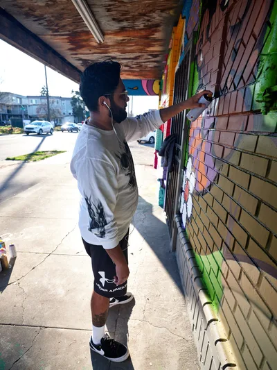 Muralist Heysus concentrates intensely as he adds precise details to a vibrant community mural on a brick wall at the intersection of Killingsworth and Northeast 15th Avenue in Portland's Alberta district. The weathered overhang casts dramatic shadows across the concrete sidewalk while afternoon light illuminates the artist's focused expression and the explosion of colors transforming the urban surface. His white long-sleeve shirt contrasts against the rainbow spectrum of spray paint emerging from his careful hand movements.