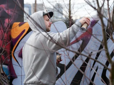 Travis Fields, known as Campographic, concentrates intently as he applies purple spray paint to a concrete wall at the Clay Creative Building in southeast Portland. The hooded artist works methodically through bare winter branches that create natural framing in the foreground, while colorful existing graffiti provides a vibrant backdrop. Soft overcast light illuminates this intimate moment of urban artistry in the Hosford-Abernethy neighborhood.