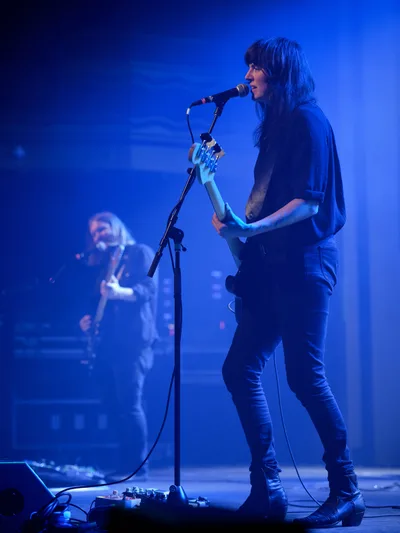 Emma Richardson of Band of Skulls commands the stage at Webster Hall during the band's 10 Year Anniversary Tour, her silhouette illuminated against a deep cobalt backdrop. The intimate venue's atmospheric lighting casts dramatic shadows as she performs alongside bandmate Russell Marsden, who remains partially visible in the misty background. Richardson's focused expression and dynamic posture capture the raw energy of live indie rock performance in Manhattan's historic East Village music venue.