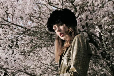 A portrait of Rhianna Atwood standing in front of a fully bloomed cherry blossom tree, its soft pink flowers filling the background. She wears an olive green crocodile-textured leather coat and a black fluffy fur hat, with long brown hair resting over her shoulders. She gazes downward with a serene expression in warm natural sunlight.