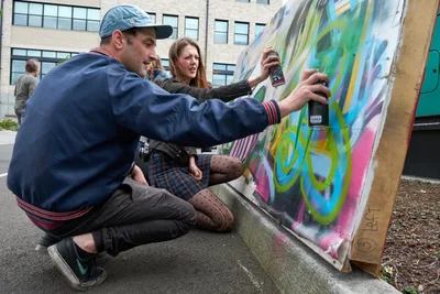 Galen Malcolm of the Portland Street Art Alliance crouches beside a vibrant canvas, spray can in hand, while demonstrating live graffiti techniques to an engaged woman in polka-dotted tights. The scene unfolds against the backdrop of Portland's Taylor Electric Blocks, where colorful street art education meets community engagement. Warm afternoon light illuminates the collaborative moment as the instructor guides his student through the rhythmic motions of aerosol artistry.