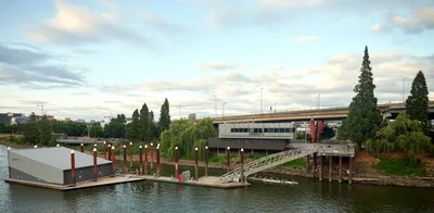 Portland Fire & Rescue Station 21 sits anchored in the Willamette River beneath the Southeast Madison Street Bridge, its modern floating structure serving the Central Eastside industrial district. The utilitarian gray building extends over dark waters on wooden pilings, connected to shore by a pedestrian gangway that spans the vegetated riverbank. Towering conifers and weeping willows frame the scene under a brooding Pacific Northwest sky, while the concrete bridge arches overhead, linking the city's urban fabric.