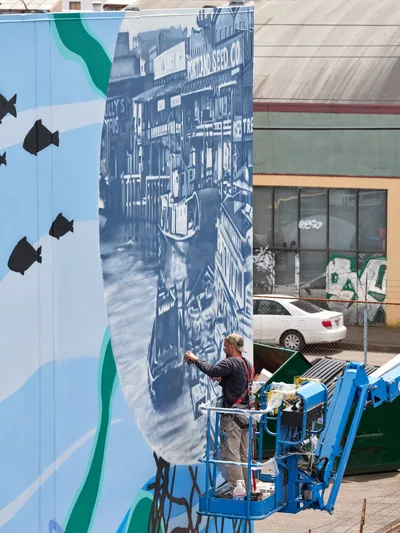 Jeremy Nichols and Devin Finley work from a blue scissor lift to paint an elaborate monochromatic riverboat mural on a building wall in Portland's Central Eastside district. The detailed artwork depicts a historic paddle steamer with visible signage including 'Natural Seed Co.', rendered in blue and white tones against the industrial urban backdrop. Black fish silhouettes swim across the adjacent wall section, creating a cohesive aquatic narrative that transforms the concrete surface into a nostalgic river scene.