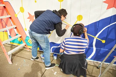 Anke Gladnick and Maria Rodriguez collaborate on a vibrant public mural titled "Let's Talk" at Open Signal in Portland's Eliot neighborhood. The standing artist in navy vest works alongside a seated partner wearing headphones and striped shirt, their brushes bringing bold geometric patterns to life against a white wall. Paint cans and a wooden ladder frame the scene as the duo transforms the community space with radiating circles and angular forms in coral, blue, and golden yellow.