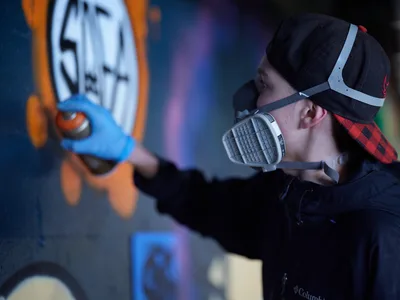In the atmospheric depths of Clay Creative's parking garage in southeast Portland's Hosford-Abernethy neighborhood, a young artist wearing a protective respirator and backwards cap applies vibrant blue paint to an emerging mural. The scene captures the intersection of urban art and industrial space, with colorful graffiti work illuminated against the concrete walls. Spray paint equipment and artistic materials suggest an active creative process unfolding in this converted creative space.