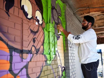 Muralist Heysus concentrates intently as he applies electric lime green paint to a dynamic street art composition on the brick wall of a building in Portland's Alberta neighborhood. The bearded artist, wearing a white hoodie and glasses, works methodically on his colorful creation featuring bold purple, orange, and pink elements that flow across the textured masonry surface. Afternoon light filters through the covered workspace, illuminating both the emerging artwork and the focused determination of the creator.