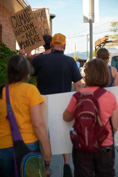 A diverse group of environmental protesters moves through McLoughlin Promenade in Oregon City, their handmade cardboard signs demanding immediate climate action rather than moderate approaches. The afternoon light bathes the small-town street scene as demonstrators in colorful casual attire—yellow shirts, orange caps, and red backpacks—walk past brick buildings and parked vehicles. The image captures grassroots activism in motion, with protesters of various ages united in their environmental advocacy along this Oregon community thoroughfare.