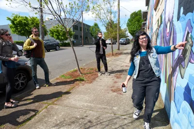 Tiffany Conklin of the Portland Street Art Alliance gestures enthusiastically toward a vibrant purple and blue mural while leading an interpretive walking tour in Portland's Central Eastside neighborhood. The afternoon light illuminates the group of engaged participants scattered along the sidewalk, some leaning against cars while others stand attentively. The scene captures the grassroots cultural energy of Southeast Portland's thriving street art community, with newly planted street trees and residential buildings forming the backdrop.