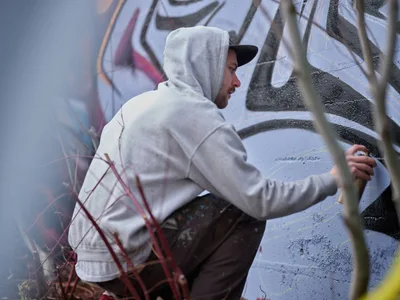 Travis Fields, known as Campographic, works intently on a large-scale mural at the Clay Creative Building in southeast Portland's Hosford-Abernethy neighborhood. Dressed in a light hoodie with hood up and black cap, the artist applies paint to the concrete wall while surrounded by urban textures and overgrown vegetation. The industrial setting and focused concentration capture the authentic spirit of Portland's thriving street art scene.