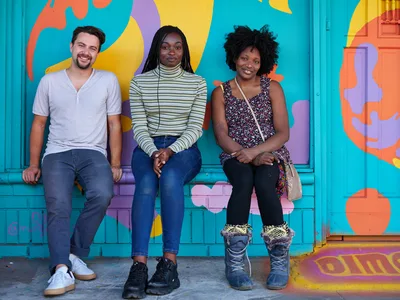 Travis Fields, Naomi Likayi, and the owner of Dime & Penny sit together against a vibrant turquoise wall adorned with flowing abstract murals in coral, yellow, and purple. The trio embodies the creative spirit of Portland's Alberta Arts District, with warm natural light illuminating their relaxed poses and genuine expressions. Their casual yet distinct personal styles—from Fields' henley and jeans to Likayi's striped turtleneck and the business owner's patterned dress—reflect the neighborhood's blend of artistic authenticity and entrepreneurial energy.