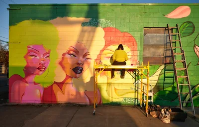 Multidisciplinary artist Villatose works from a yellow scissor lift, adding finishing touches to 'Jardín Cubana,' a stunning large-scale mural featuring three female figures with voluminous natural hair rendered in brilliant yellows, pinks, and corals. The vibrant street art transforms a green concrete block wall on Northeast Alberta Street in Portland's celebrated Alberta Arts District, where golden afternoon light bathes both the artist and their colorful creation. Painting supplies and equipment rest at the base of the wall, capturing the authentic working process of contemporary urban muralism.