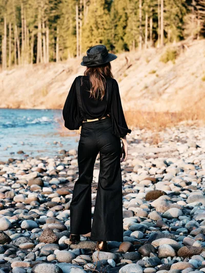 A lone woman in flowing black attire stands with her back to the viewer on the rocky shoreline of the Columbia River along Oregon's Floodplain Trail. Her wide-brimmed hat and billowing sleeves create dramatic silhouettes against the warm afternoon light that bathes the river stones and towering forest beyond. The scene captures a moment of quiet contemplation, with smooth stones of the riverbed stretching toward gentle waters while dense Pacific Northwest woodland rises from the opposite bank.