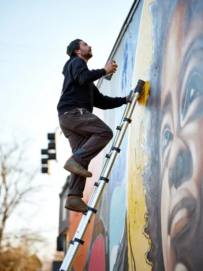 Street artist Travis Fields, working under the moniker campographic, balances precariously on an aluminum ladder as he spray paints details of poet Amanda Gorman's portrait on a building wall at Northeast 15th and Killingsworth in Portland's Alberta district. The bearded artist in dark clothing focuses intently on his work against the backdrop of pastel blues, warm oranges, and golden yellows that form the emerging mural. Urban buildings blur softly in the background under overcast Pacific Northwest skies, capturing a moment of artistic creation transforming the neighborhood streetscape.