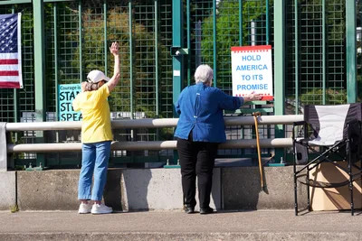 Two senior activists position protest signs along a concrete barrier overlooking Interstate 5 in North Portland's Humboldt neighborhood. One person in a bright yellow shirt waves to passing traffic while another in a denim jacket steadies a white and red sign reading "HOLD AMERICA TO ITS PROMISE." The green mesh security fencing and urban infrastructure frame this grassroots demonstration under natural daylight, with an American flag visible in the background.