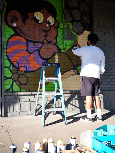 Muralist Heysus works methodically on a colorful character-based mural along the brick wall of a building in Portland's Alberta neighborhood. The artist, dressed casually in a white long-sleeve shirt and gray shorts, stands on a blue Werner ladder surrounded by an array of spray paint cans scattered across the concrete sidewalk. His emerging artwork features a whimsical monkey character rendered in warm earth tones against brilliant lime green backgrounds, capturing the dynamic spirit of Northeast Portland's thriving street art scene.