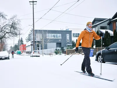 A man cross-country skis down a snow-covered residential street in Portland's Alberta Arts District during a winter snowstorm. He wears an orange jacket, black ski pants, a mustard yellow beanie, and mirrored sunglasses, smiling as he glides past parked cars. A second skier in a red jacket is visible further down the street in the background, along with modern homes, utility poles, and lightly falling snow.